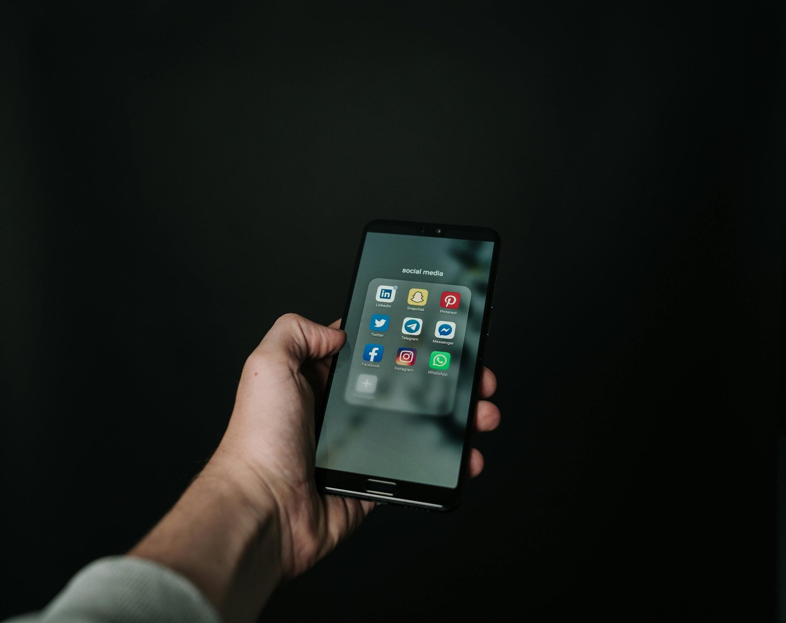 Close-up of a hand holding a smartphone displaying various social media app icons on a dark background.